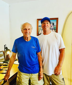 Two men standing indoors with a framed picture on the wall behind them.
