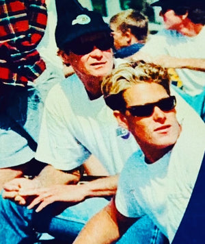 Two men sitting together, one wearing sunglasses, with a blurred background