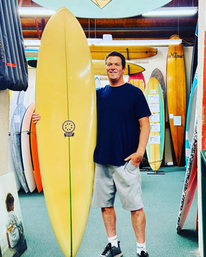 Man holding a yellow surfboard in a store with various surfboards in the background