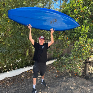Man holding a blue kayak above his head outdoors with trees and a wooden fence in the background.