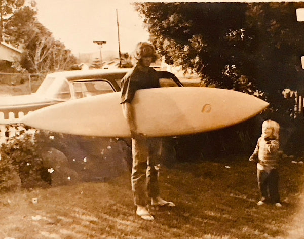 Vintage photo of a person holding a surfboard with a child standing next to them, outdoors.