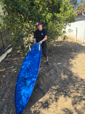 Person holding a blue surfboard on a concrete surface with trees in the background