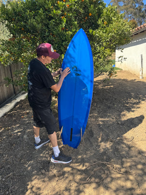 Person holding a blue kayak on a dirt path with trees and a house in the background